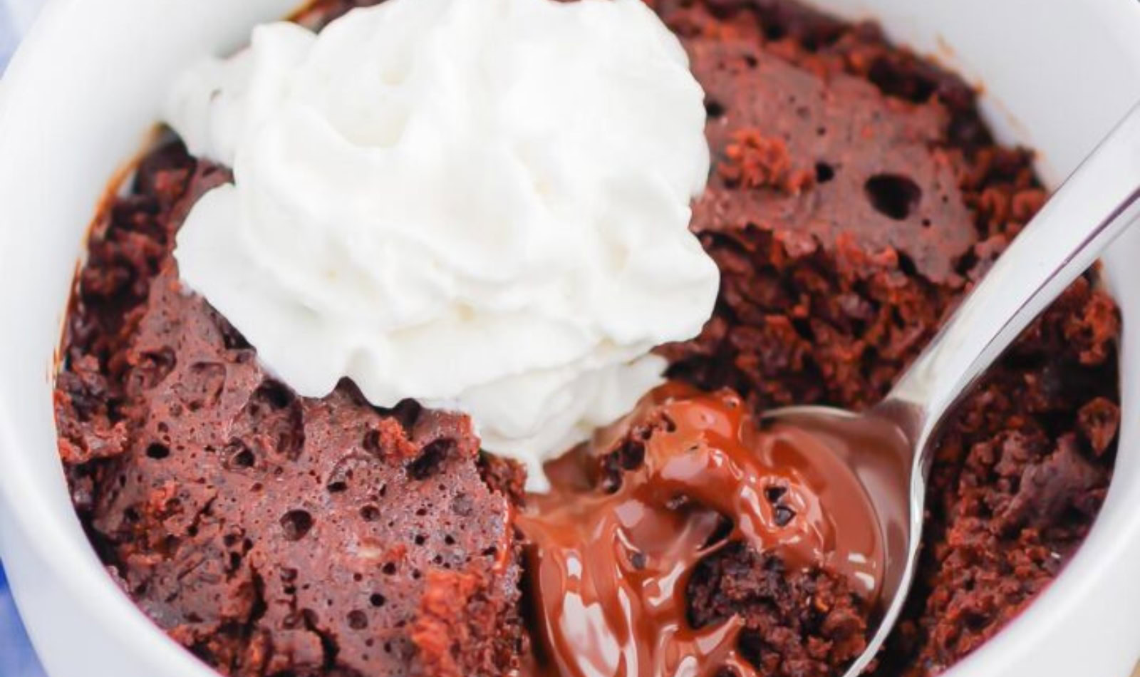 image of Chocolate-Lava-Mug-Cake with whipped cream on top all in a white mug with a silver spoon holding a bite of chocolate lava