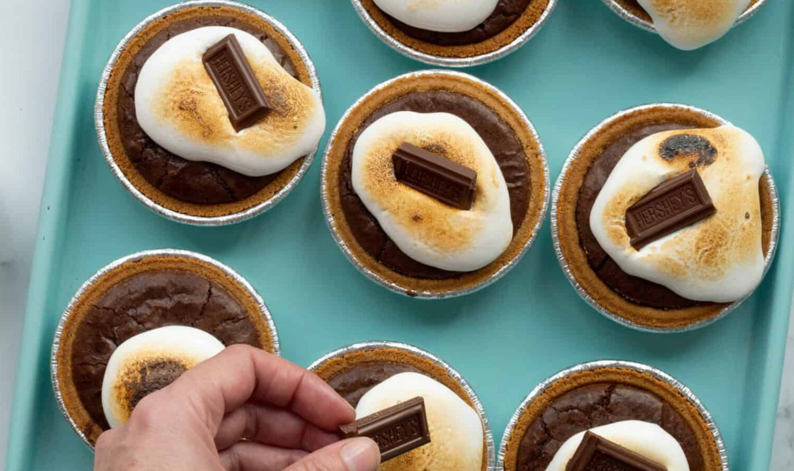 Image of mini smores pies with a hand putting a chocolate piece on top. 