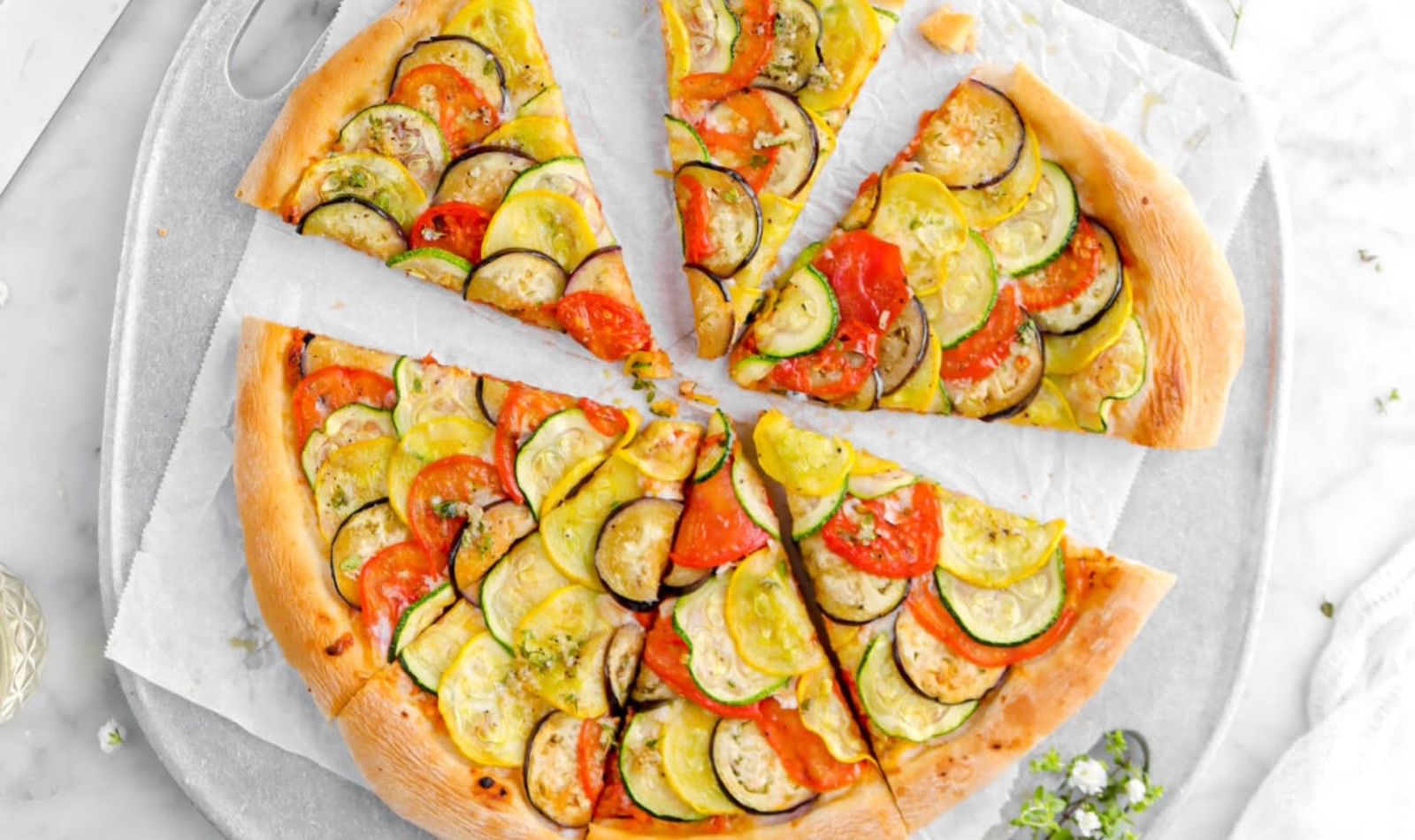 Image of a pizza on a white plate on a marble tabletop. Pizza has tomatoes summer squash and zucchini