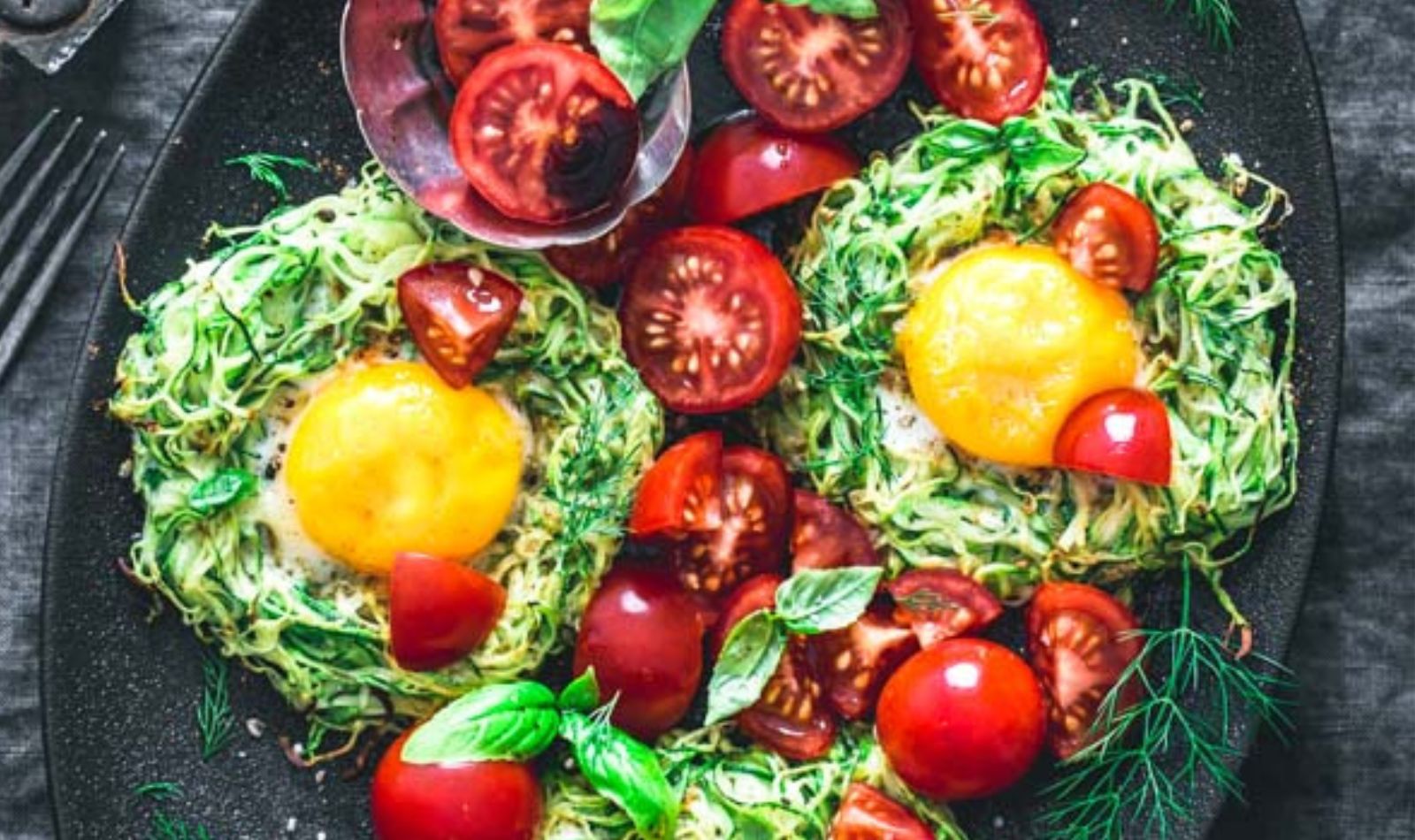 Image of zucchini egg nests made of spiralized zucchii with egg on top and garnished with sliced tomatoes and herb.